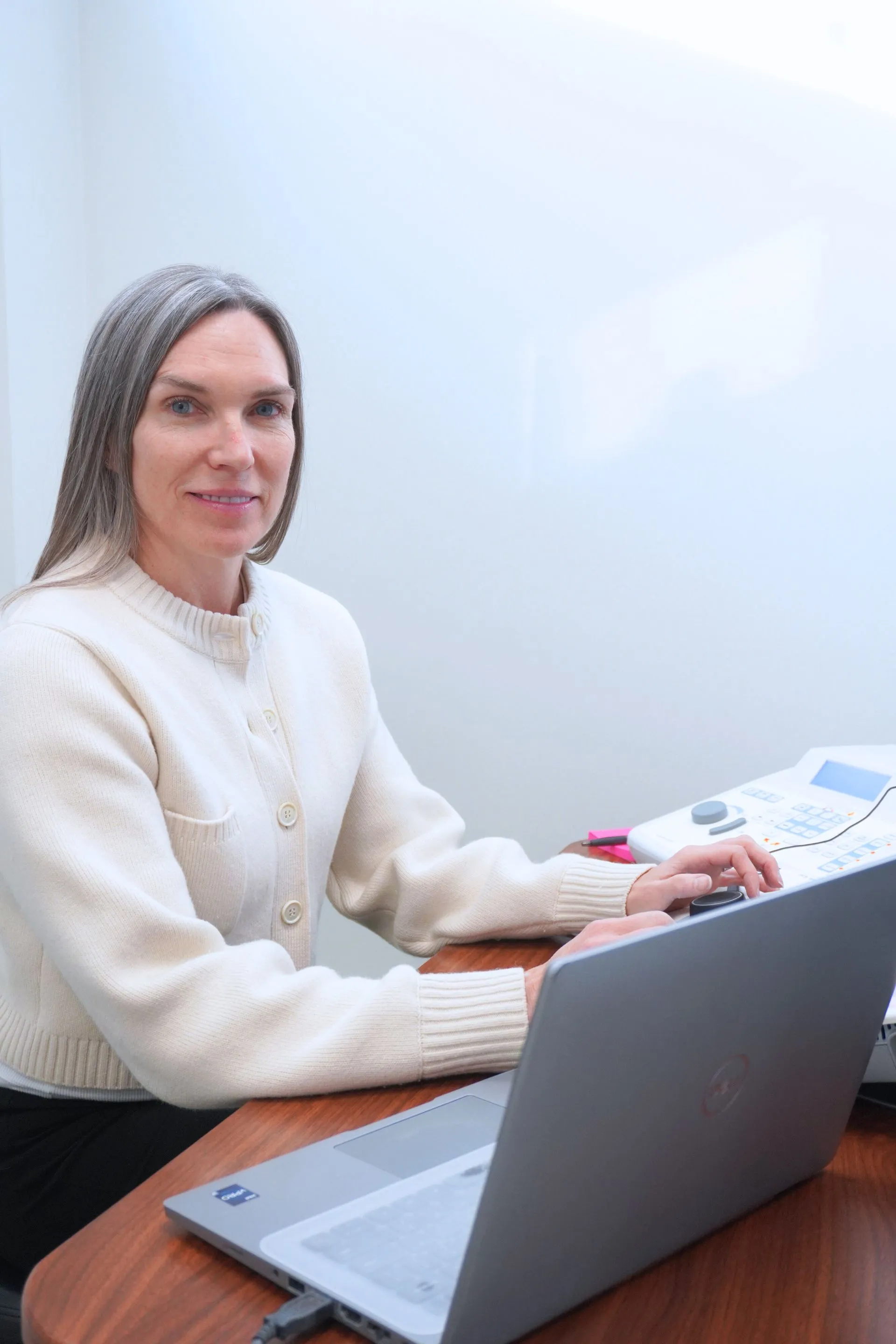 A person sits at a wooden desk with a laptop and a medical device in a brightly lit room, looking toward the camera.