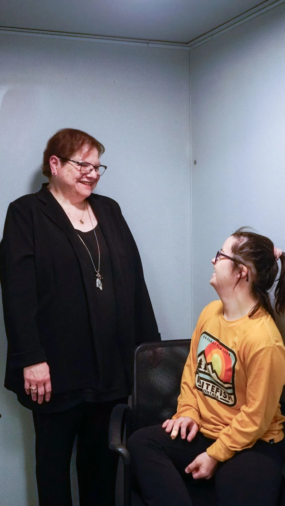 A person in a black blazer stands and smiles down at someone seated in a yellow long-sleeved shirt in a light-walled room.