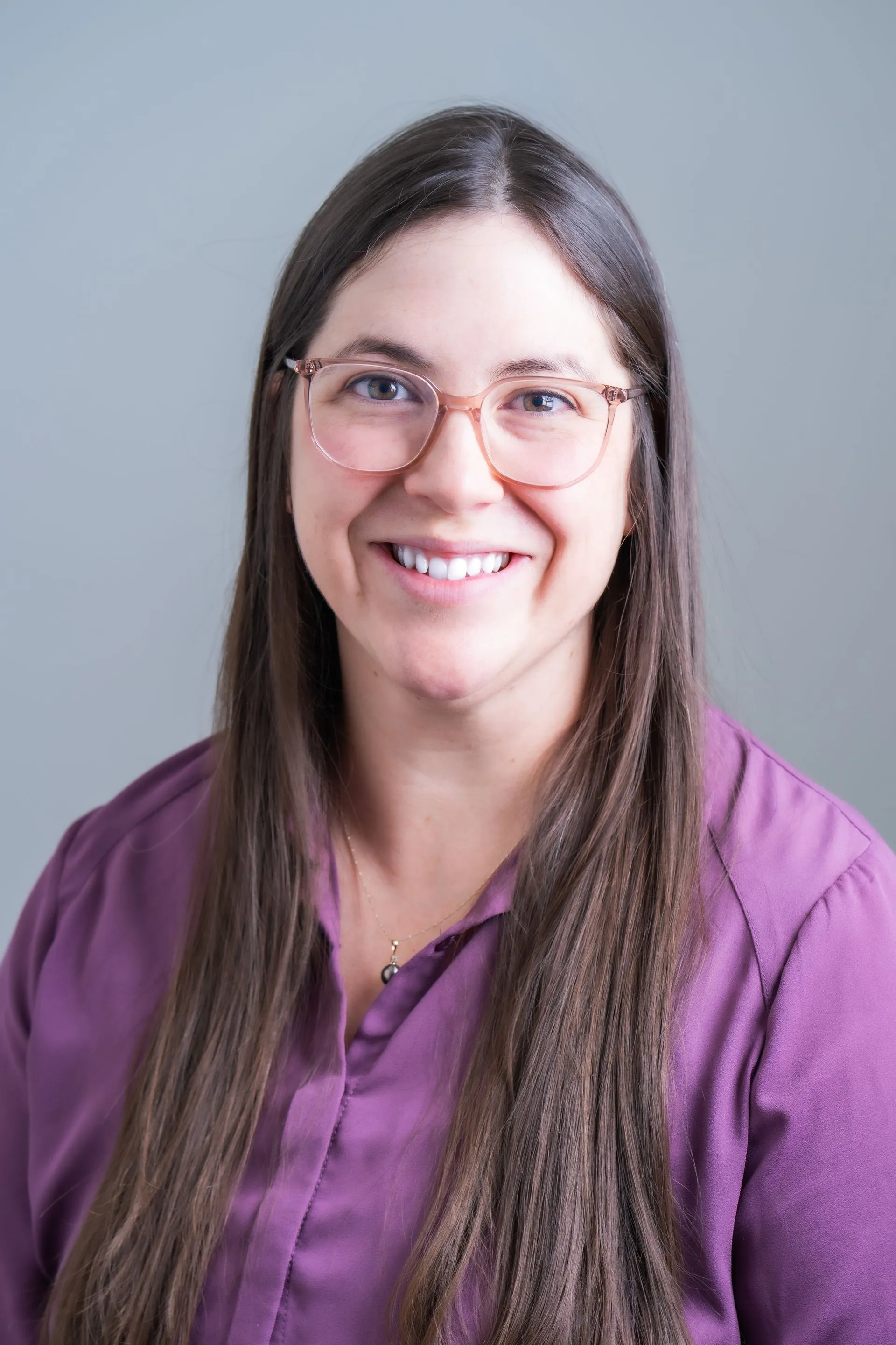 A person with long brown hair and glasses wearing a purple collared shirt, smiling against a plain gray background.