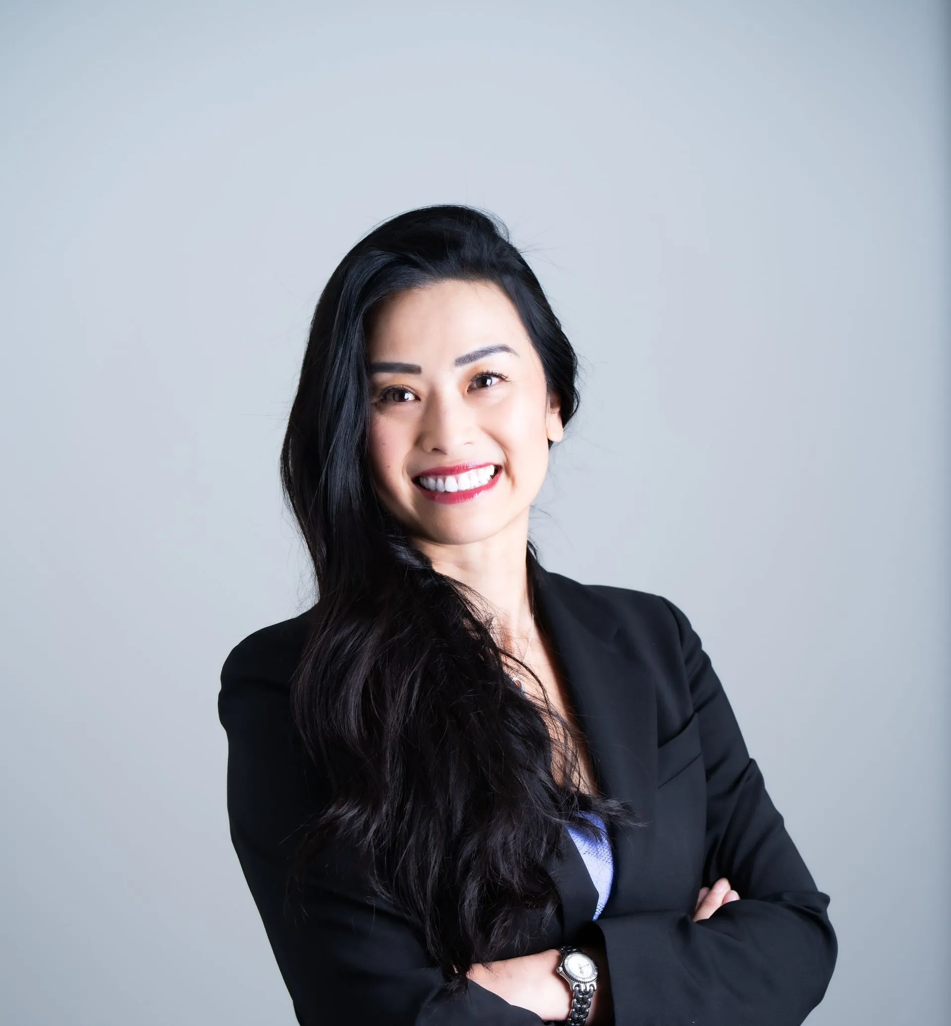 A person with long dark hair, wearing a black blazer, smiles with arms crossed against a light grey background.