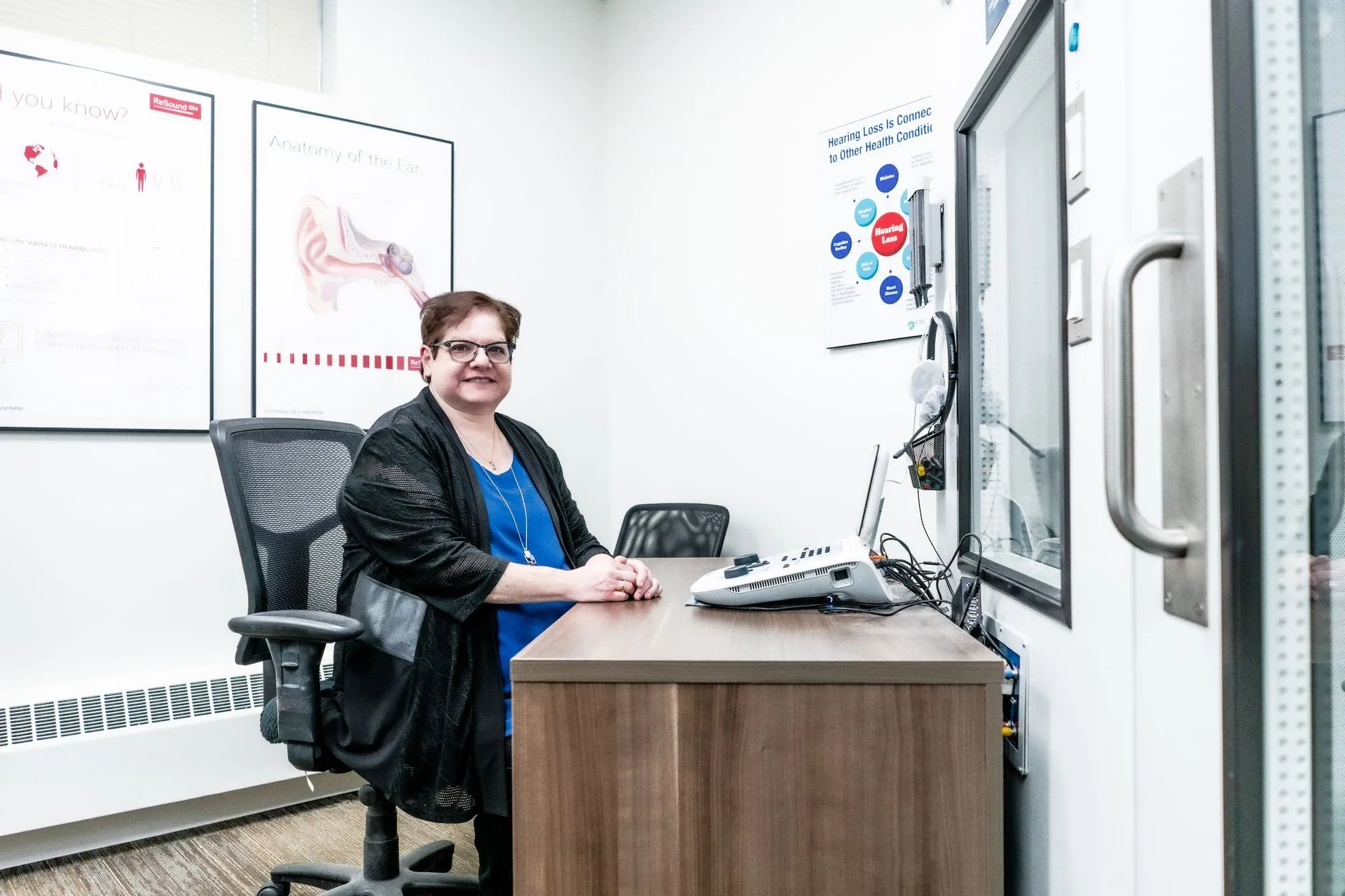 A person sits at a wooden desk with an audiometer in a medical office, with anatomical ear diagrams on the wall.