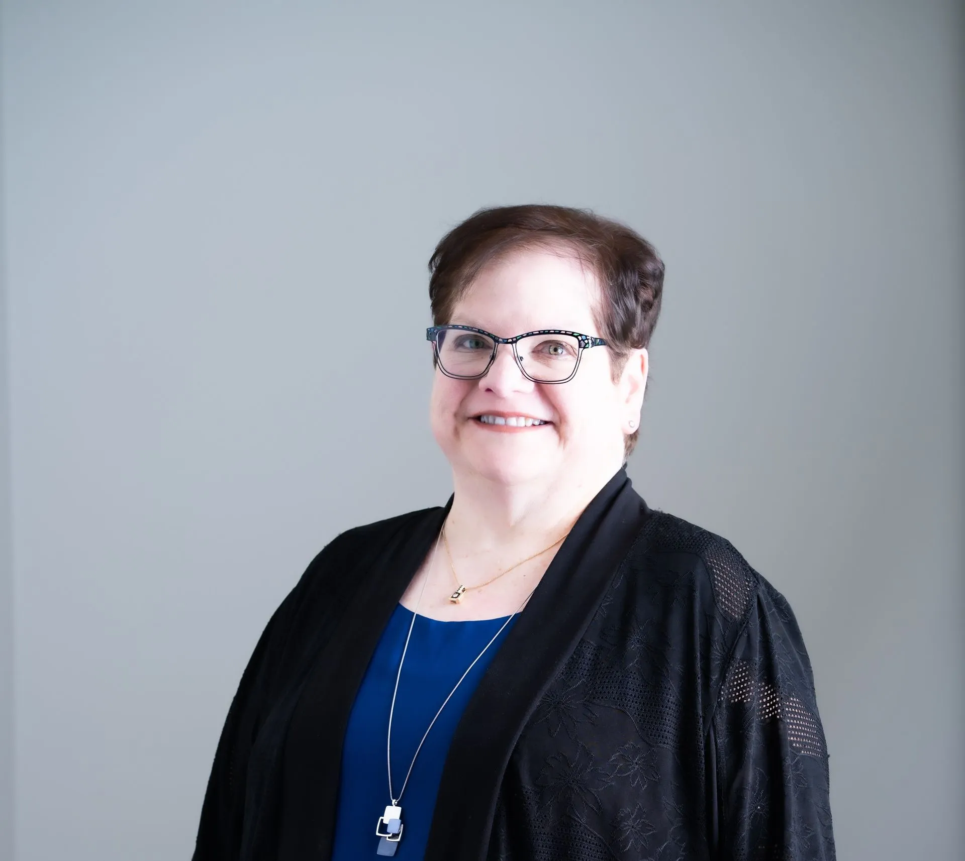 A person with short brown hair wearing glasses, a blue top, and a black cardigan smiles against a plain gray background.
