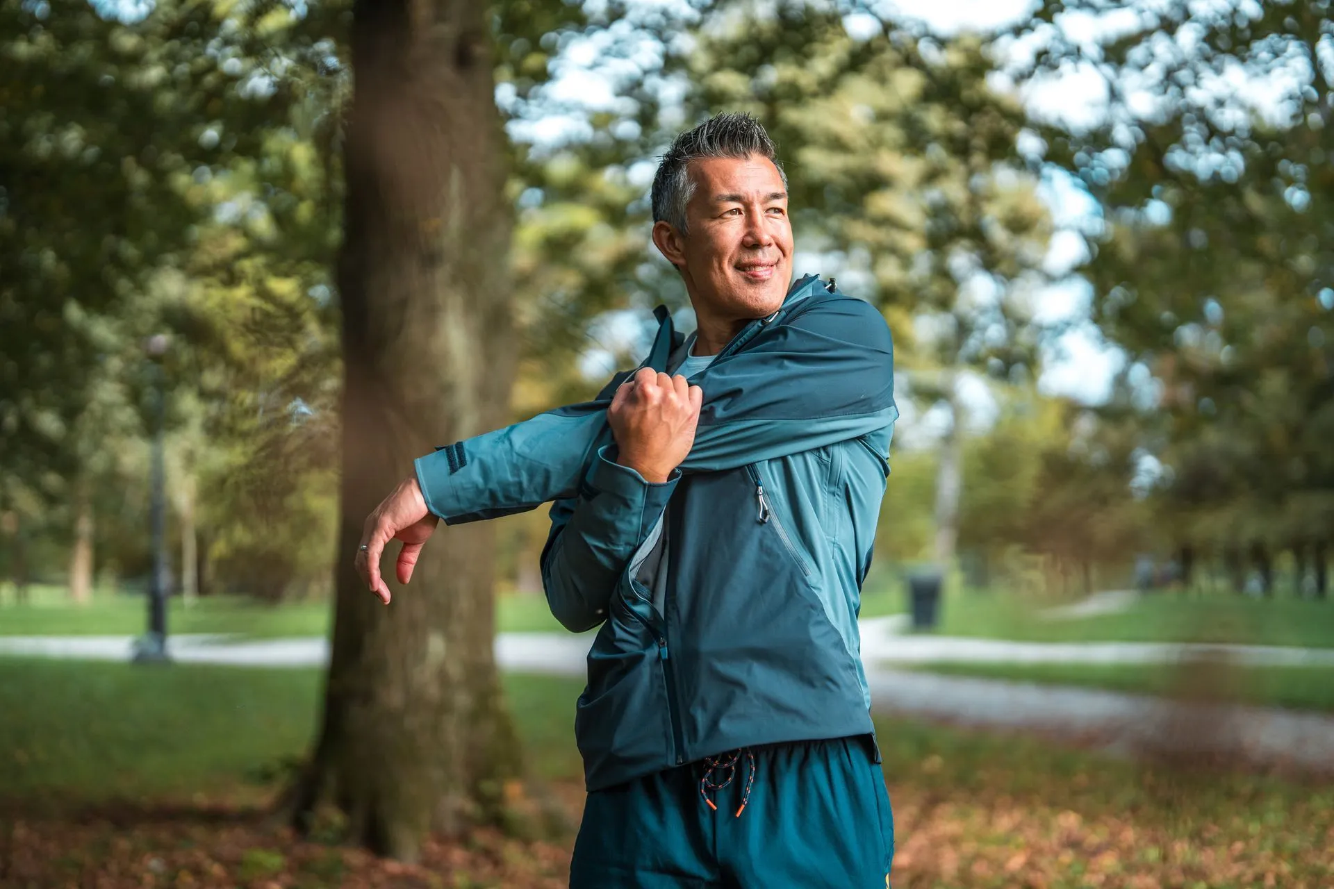 A person in a blue windbreaker and shorts stretches their arm across their chest while standing in a park.