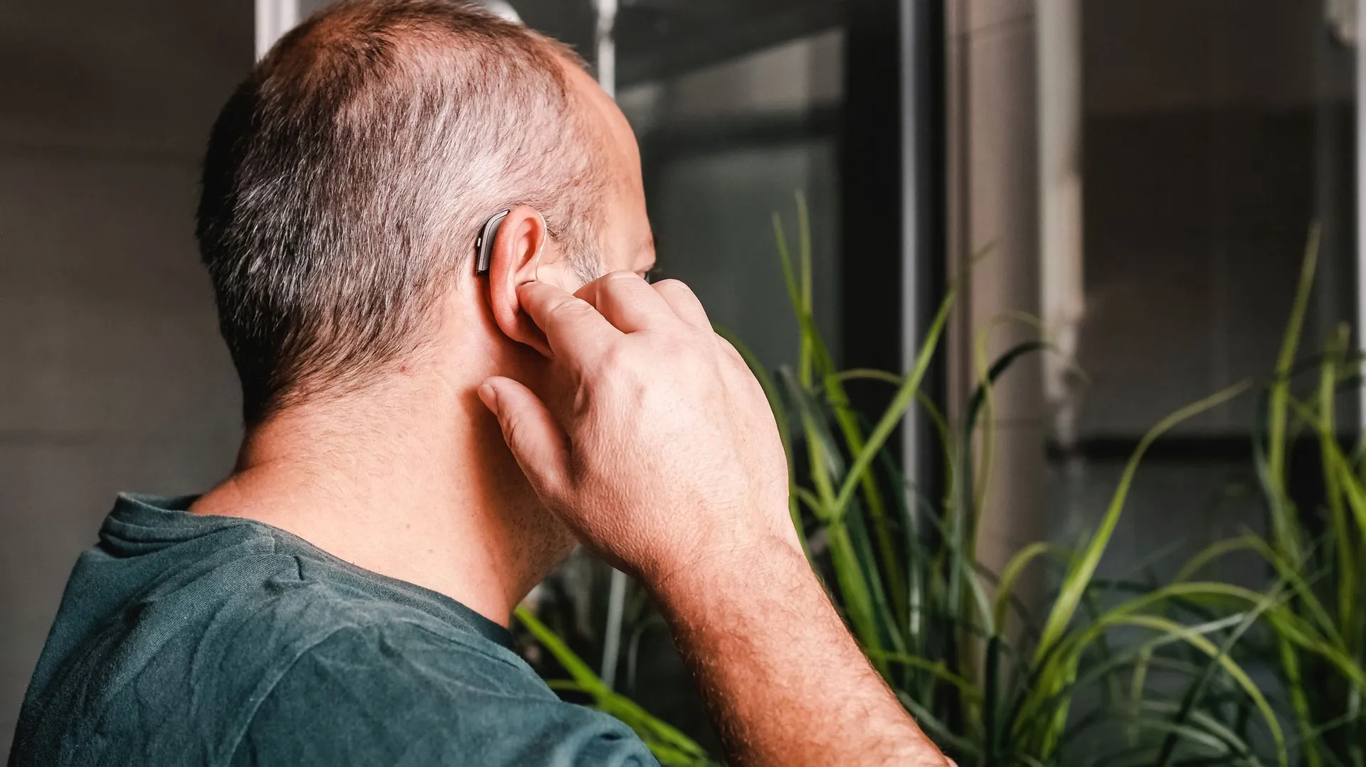 A person adjusting a behind-the-ear hearing aid while standing near a houseplant.