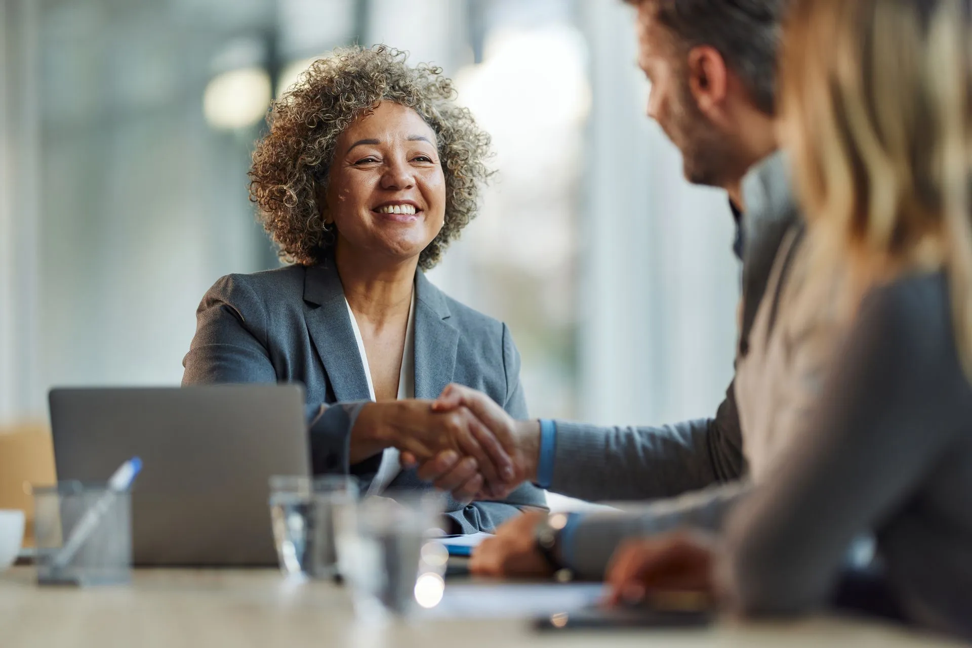 A professional smiling at colleagues while shaking hands across a desk in an office setting with a laptop nearby.