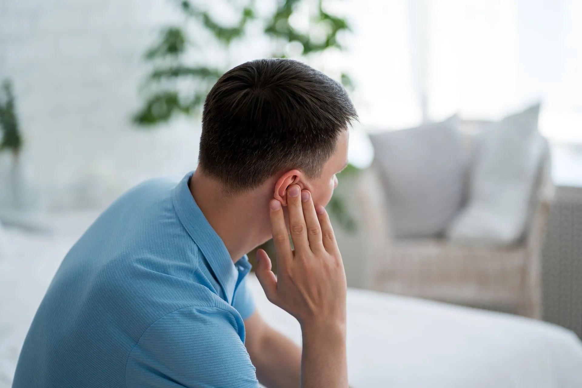 A person in a blue shirt sitting on a bed, holding their ear, appearing to experience discomfort.