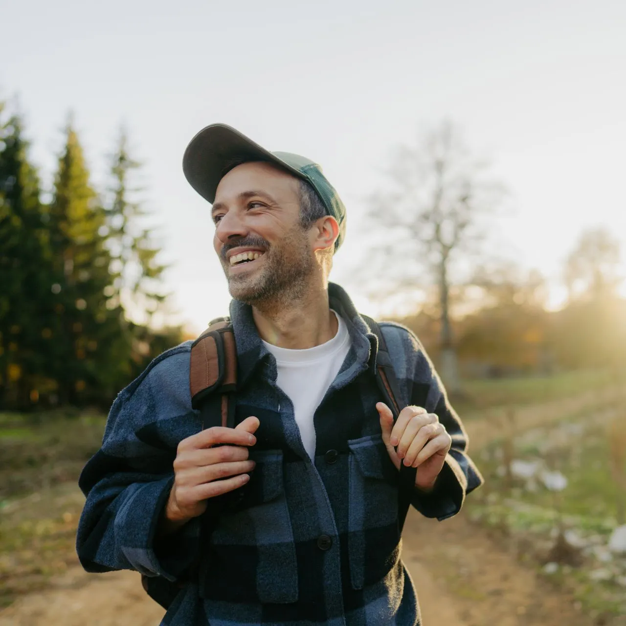A smiling person in a plaid shirt and baseball cap wearing a backpack, walking along a dirt path in a forest at sunset.