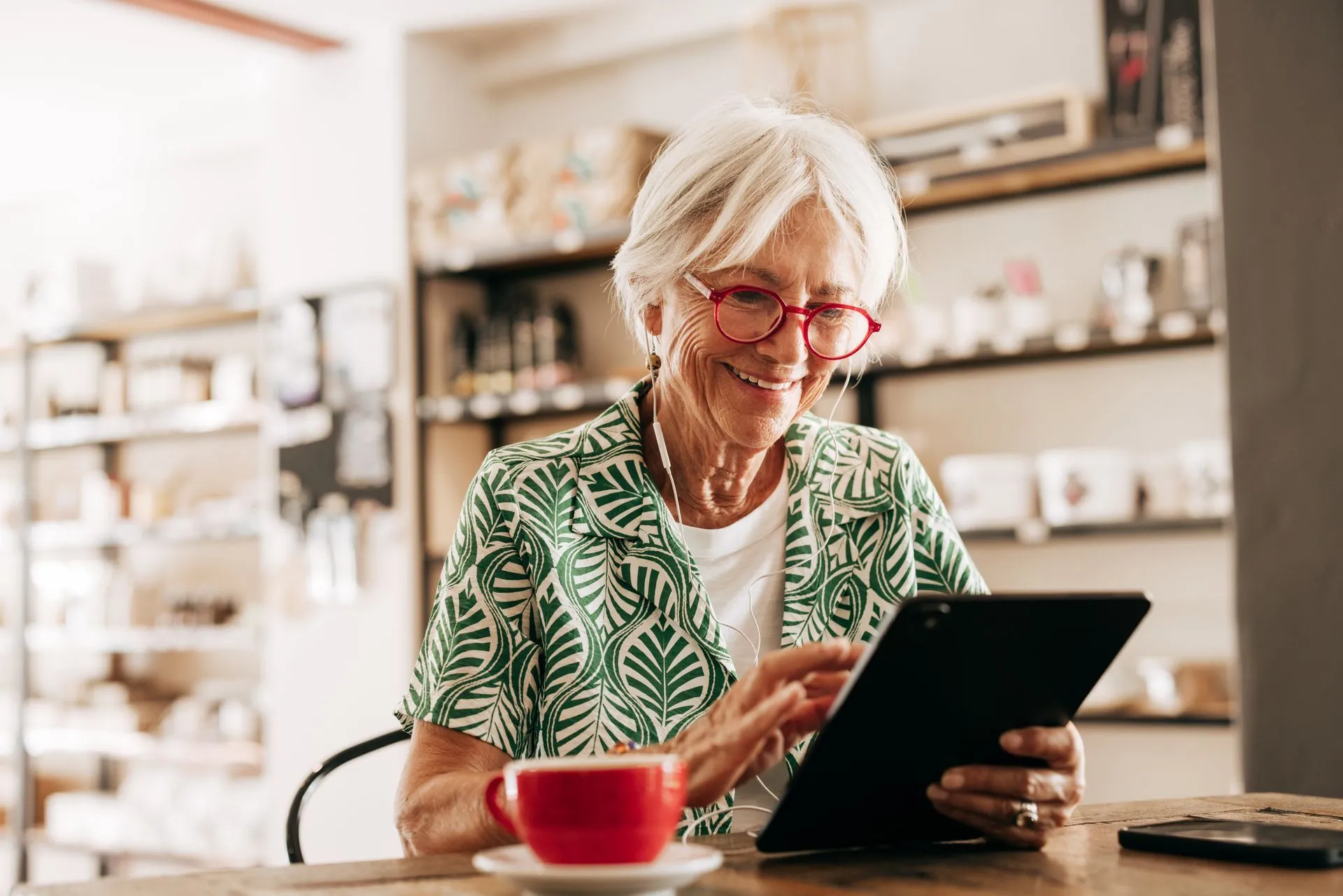 A person in a green leaf-print shirt smiling while using a tablet at a table with a red cup of coffee.