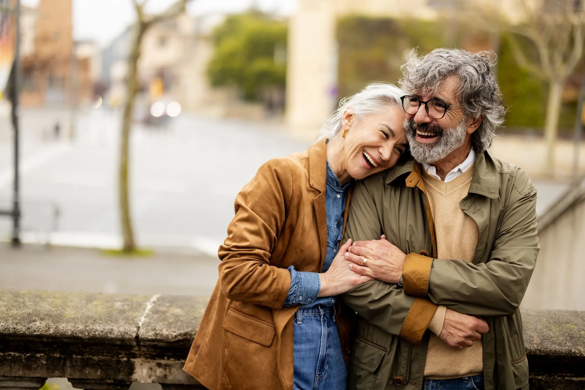A couple standing together outdoors, smiling and laughing while leaning on each other against a stone wall.